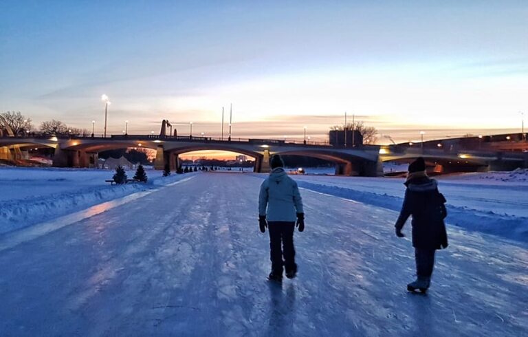 People walking on a frozen Winnipeg river trail under a bridge at winter sunset