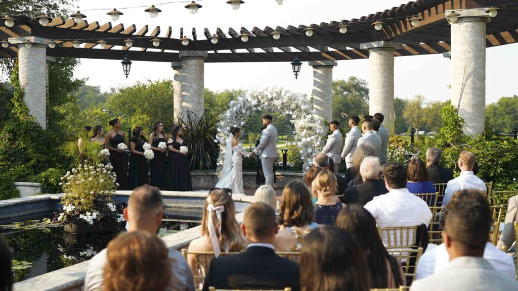 Outdoor wedding ceremony with couple exchanging vows under a garden pergola in front of guests