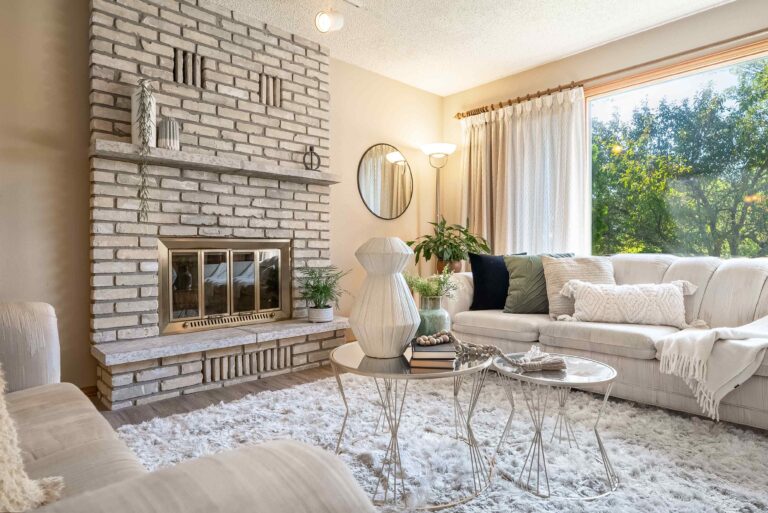 Staged living room with brick fireplace, white sofa, large window, and natural light